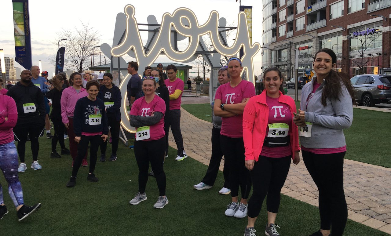 Group of runners wearing pink Pi t-shirts gathered under a public sculpture that is a large image of the word "joy"