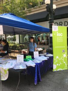 Tables on street displaying t-shirts and science signage with blue canopy and 2 women smiling standing beside Illinois Science Council green banner