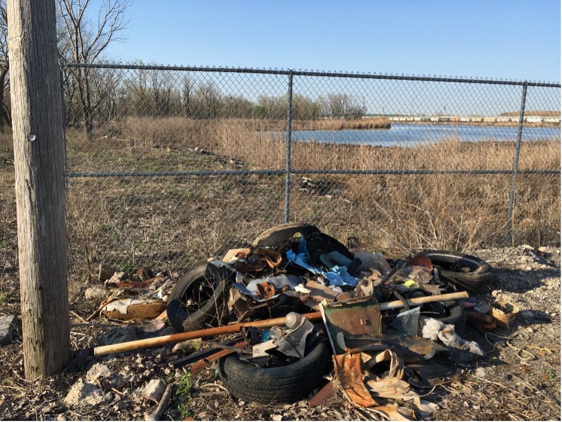 Photo of pile of garbage in front of chain link fence in field with brown grass with water in background.